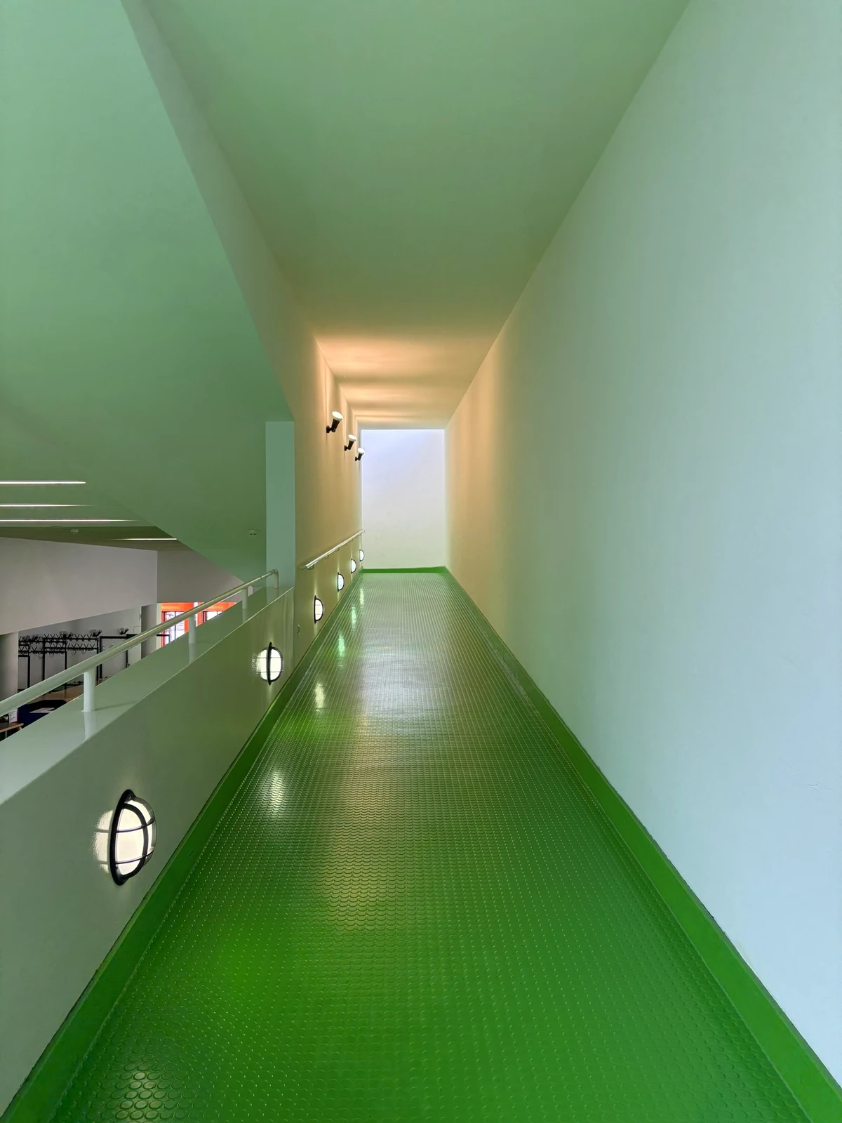 A green-floored walkway with pale walls and small round lamps